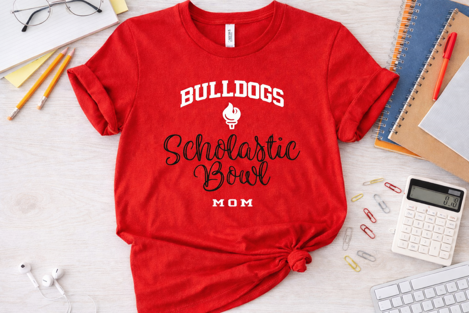 Red t-shirt with 'Bulldogs Scholastic Bowl Mom' text on a desk with office supplies.