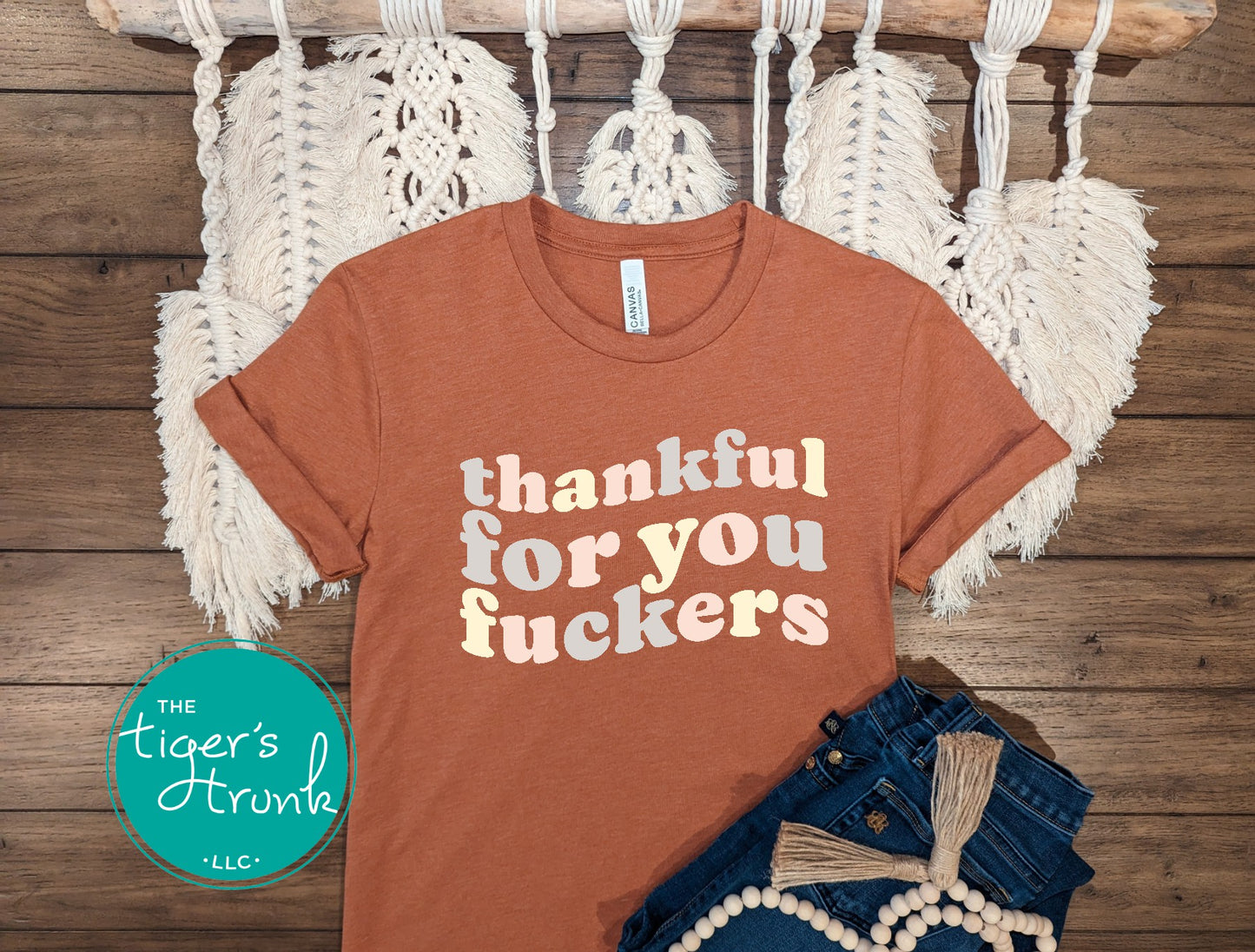 Brown t-shirt with 'thankful for you fuckers' text on a wooden surface with macrame hanging.
