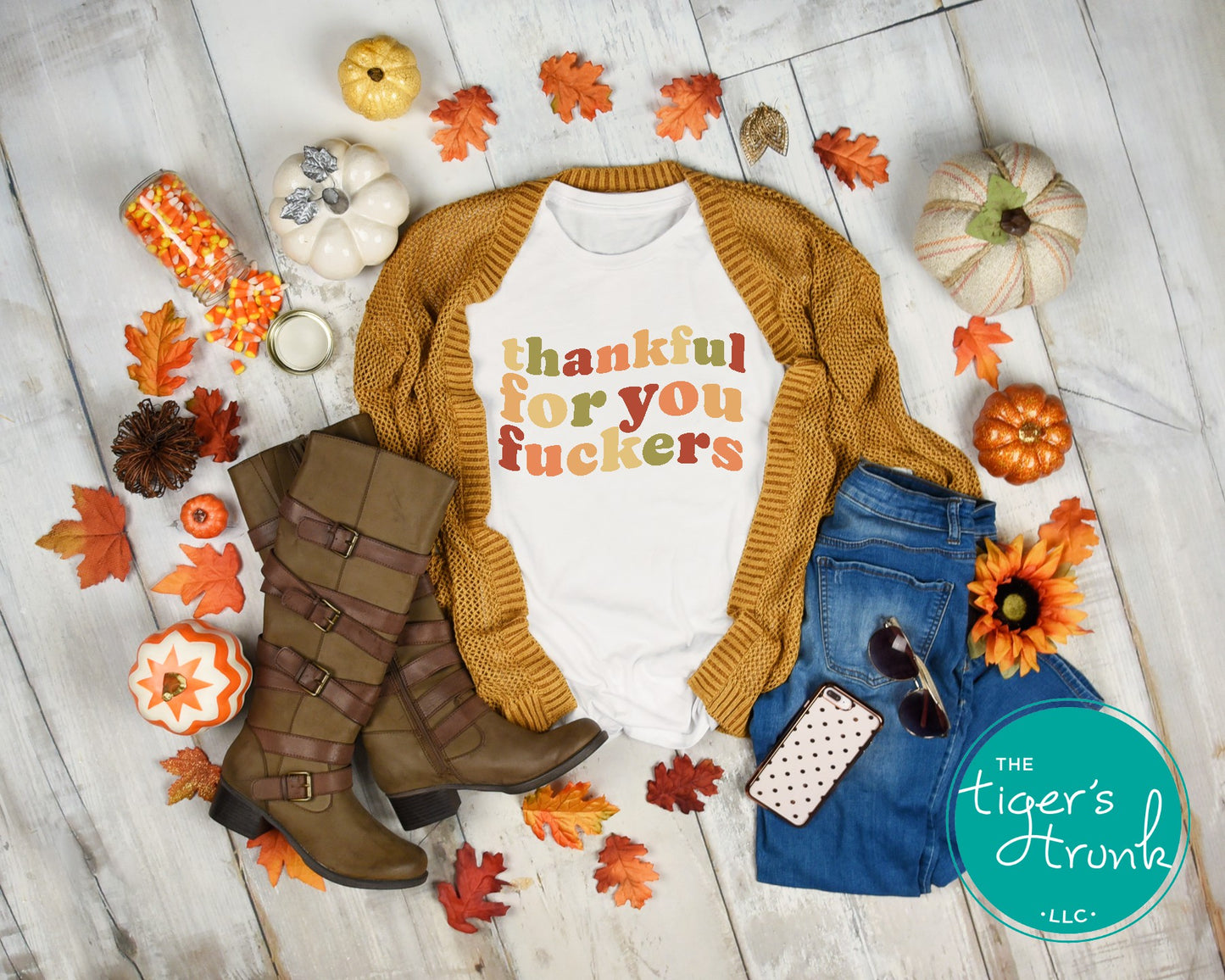 Fall-themed outfit with sweater, jeans, boots, and 'thankful for you fuckers' shirt on a wooden floor with pumpkins and leaves.