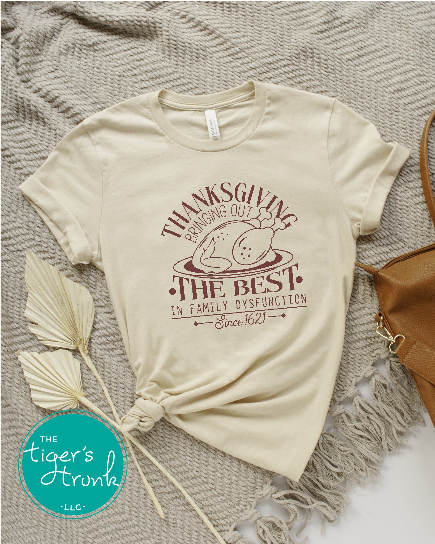 A beige short-sleeve thanksgiving themed shirt laid out flat on a grey textured surface with a text graphic and some decorative feathers. The shirt has a printed text that reads 'Thanksgiving: Bringing Out the Best in Family Dysfunction Since 1621'.