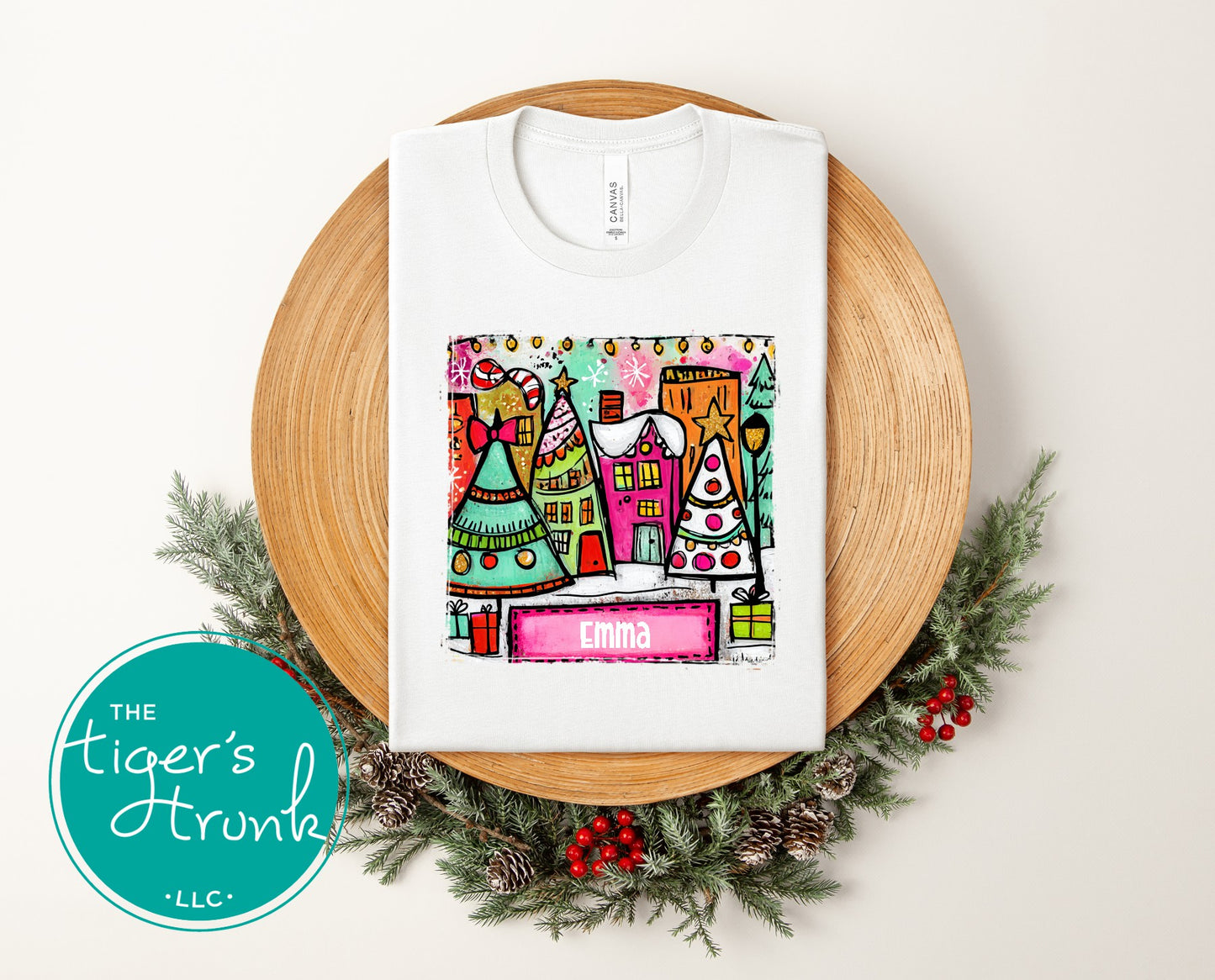 White t-shirt with colorful Christmas village design on a wooden plate with greenery, featuring 'The Tiger's Den' logo.