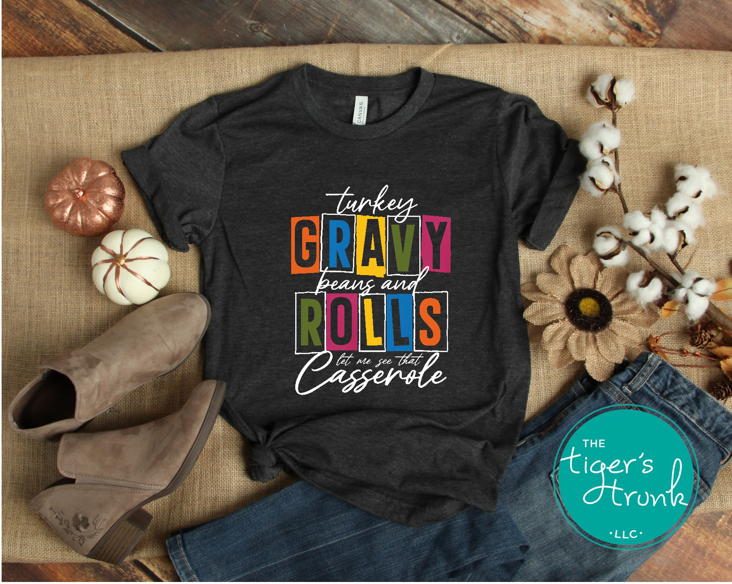 A black short-sleeve Thanksgiving themed shirt with text 'Turkey, Gravy, Beans and Rolls, Let Me See That Casserole' printed on it, displayed flat with a pair of shoes and some decorative items around it.