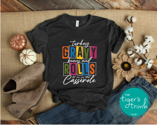A black short-sleeve Thanksgiving themed shirt with text 'Turkey, Gravy, Beans and Rolls, Let Me See That Casserole' printed on it, displayed flat with a pair of shoes and some decorative items around it.