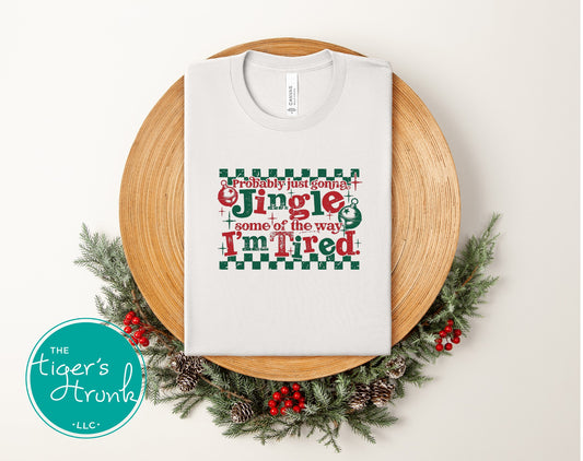 White t-shirt with Christmas-themed text on a wooden plate surrounded by decorative elements.