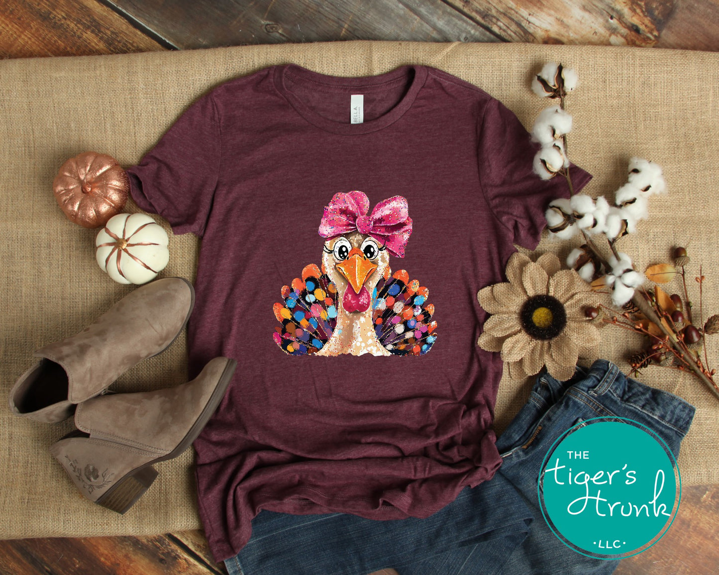 Maroon t-shirt with a turkey graphic and bow, surrounded by fall-themed items on a wooden surface.