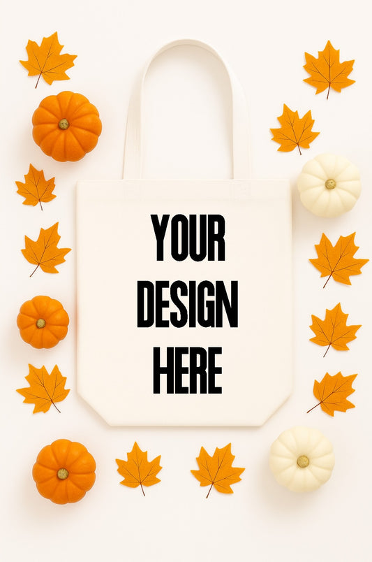 White tote bag with 'Your Design Here' text, surrounded by pumpkins and autumn leaves on a light background