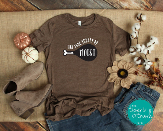 A brown short-sleeve Thanksgiving themed shirt with the text 'MAY YOUR TURKEY BE MOIST' printed on the front, displayed flat with a pair of blue jeans and a pair of brown boots on either side, placed on a wooden surface with decorative flowers and a toy dog in the background.
