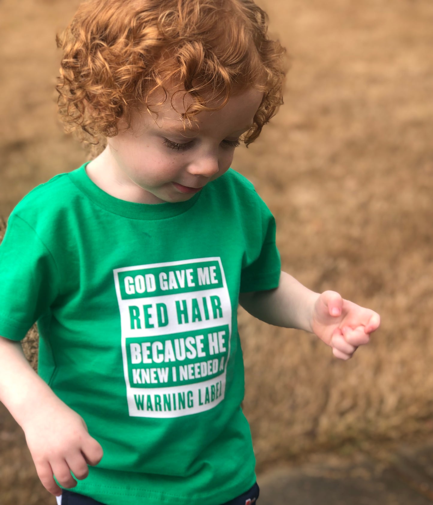 Young child wearing green St. Patrick’s Day red hair warning label shirt, looking down.