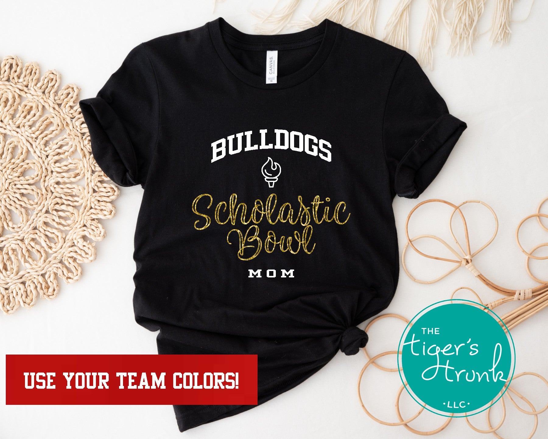 A black short-sleeve t-shirt with 'Bulldogs' and 'Scholastic Bowl Mom' text in white and gold colors, displayed on a surface with a white circular decoration and a beige fan behind it.