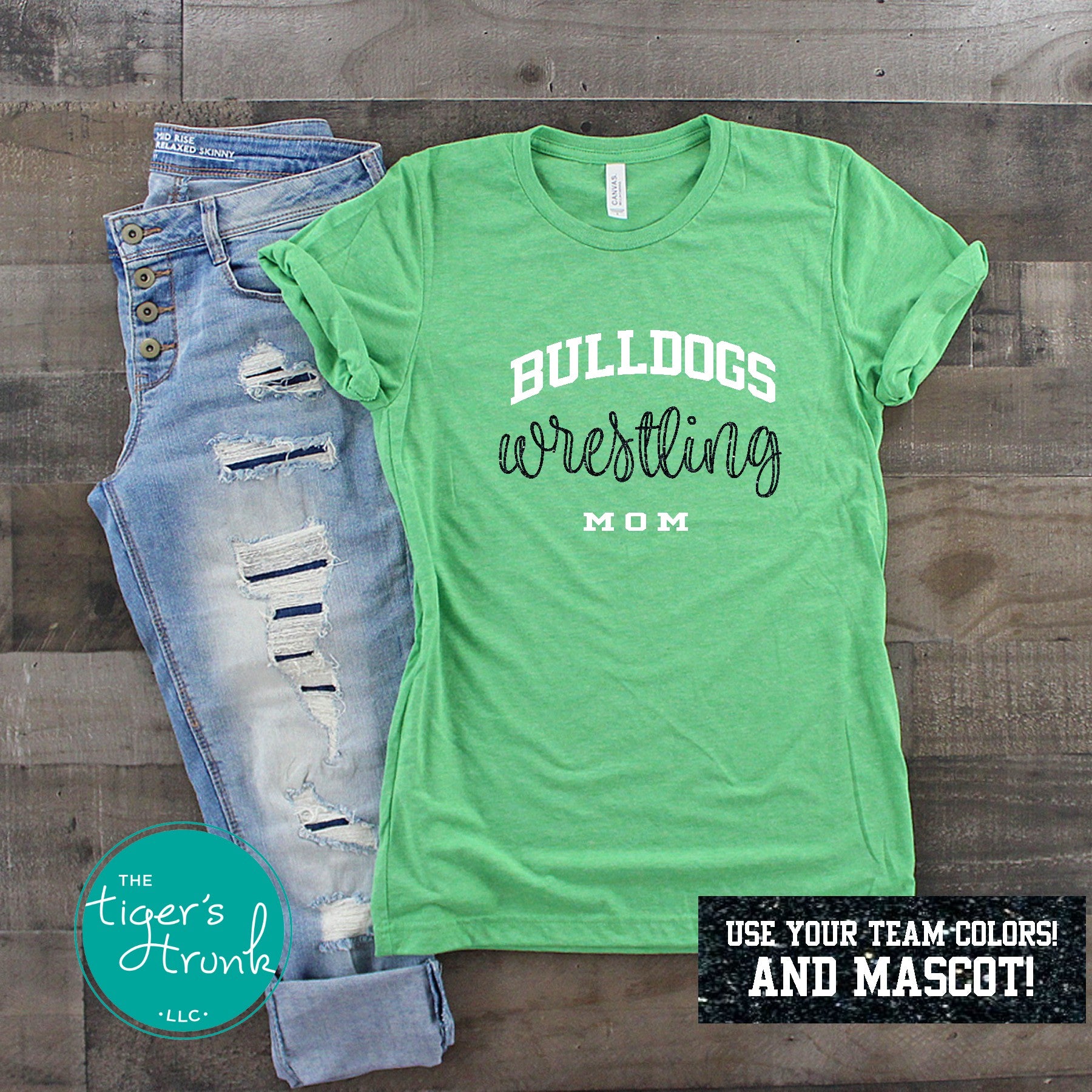 A short-sleeve green shirt with the text 'Wrestling Mom' and a school mascot, paired with blue jeans. The shirt is displayed on a wooden surface with a text overlay indicating that it can be customized with a team mascot and colors.