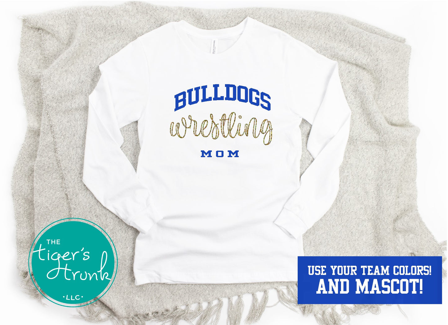 A white long-sleeve wrestling shirt with 'WRESTLING MOM' printed in blue and gold, displayed on a grey blanket with a logo at the bottom.
