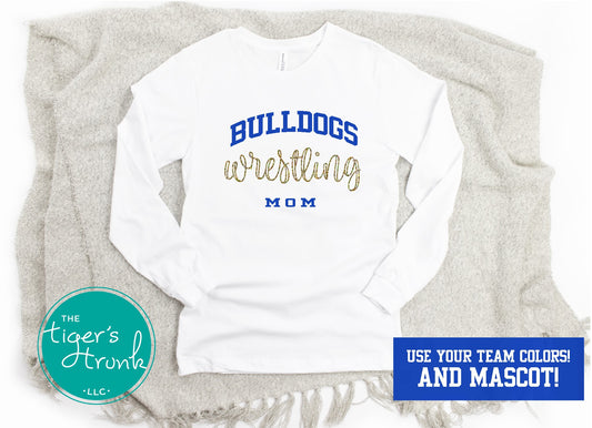A white long-sleeve wrestling shirt with 'WRESTLING MOM' printed in blue and gold, displayed on a grey blanket with a logo at the bottom.